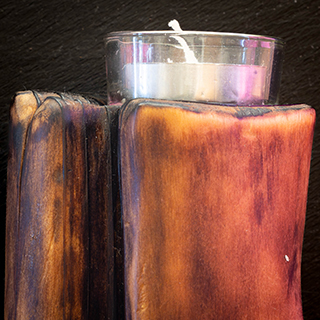 A tight, atmospheric close-up shot of a glowing tea light nestled within the gnarled, scorched texture of a hand-burnished tree branch holder.