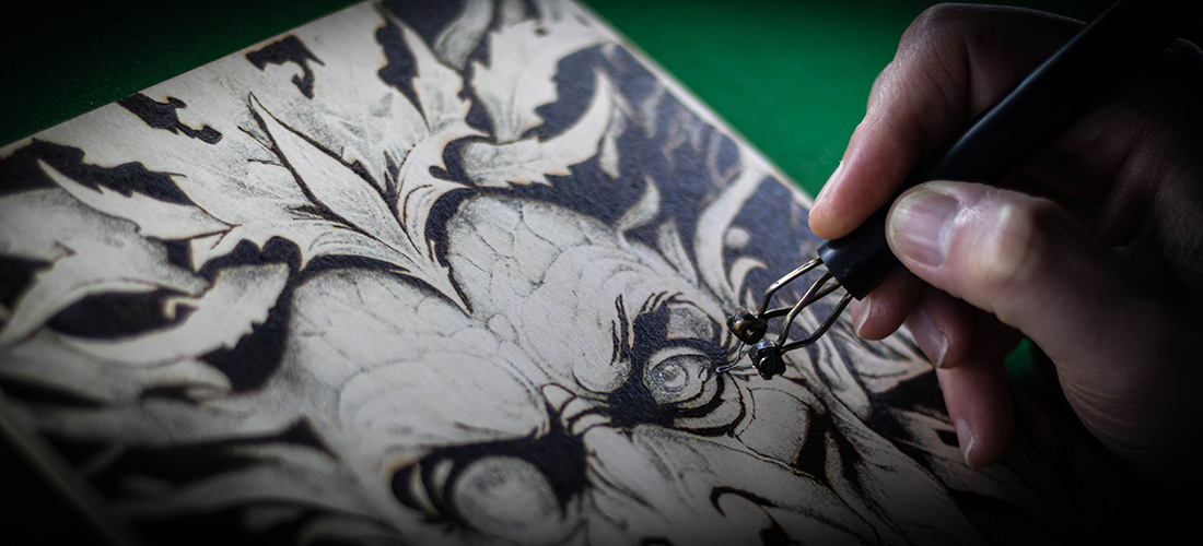 Macro action shot of a hand using a pyrography pen to burn a Green Man design into FSC Birch Plywood. The metal nib is in sharp focus against the wood grain, highlighting the heat and precision of the wood-burning craft.