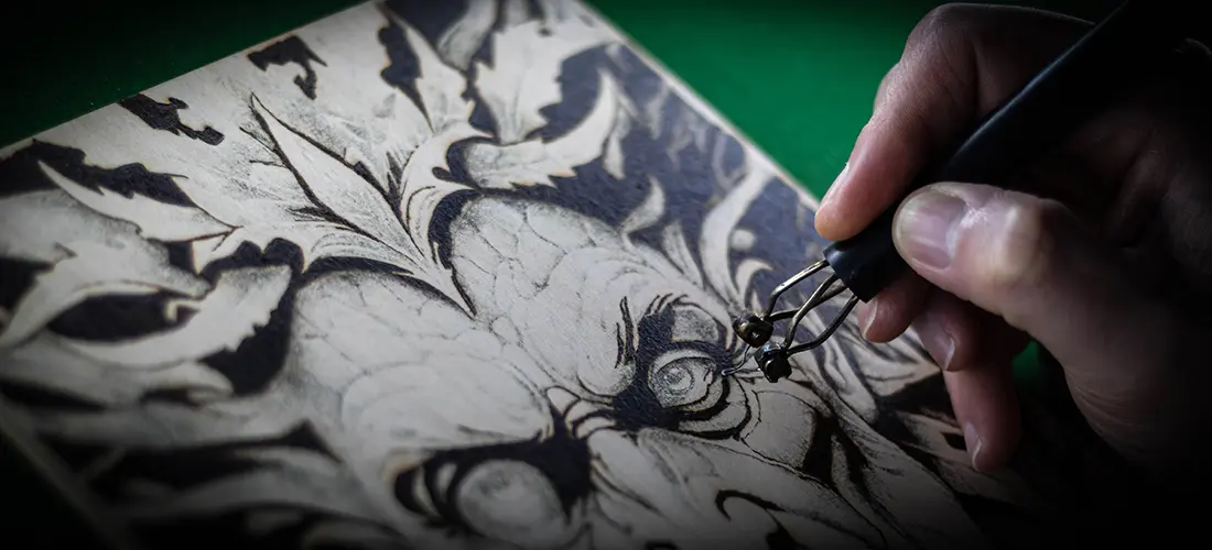 Macro action shot of a hand using a pyrography pen to burn a Green Man design into FSC Birch Plywood. The metal nib is in sharp focus against the wood grain, highlighting the heat and precision of the wood-burning craft.