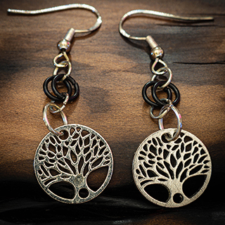 A pair of silver-toned chainmail drop earrings displayed against the dark, textured background of a burnt wooden branch.