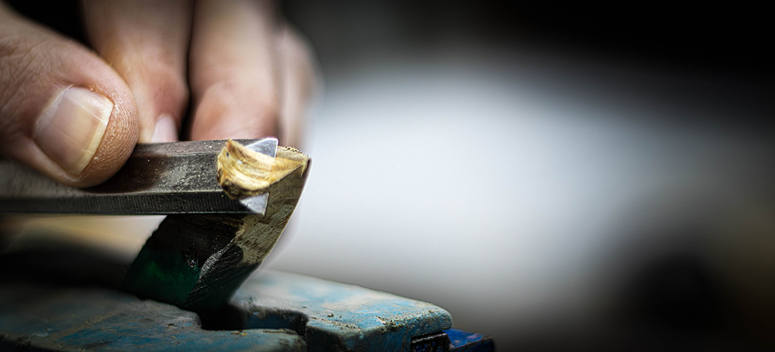A macro action shot of a hand using a chisel to carve into a block of wood, with a sharp curled shaving of timber rising from the blade against a dark, moody background.