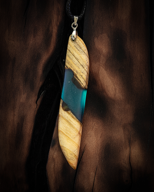 A close-up of a fused wood and translucent sea-green resin pendant resting on a gnarled, scorched wooden branch background, highlighted by soft rim-lighting.