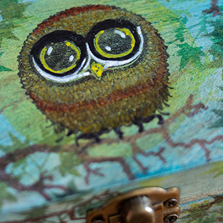 A close-up of a hand-painted owl on a wooden box, showing the brushwork and paint texture over natural wood grain.